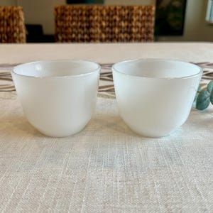 May include: Two white, opaque glass bowls with rounded bottoms and slightly flared rims. The bowls are set on a light-colored tablecloth with a subtle pattern. The background includes a woven chair and decorative elements.