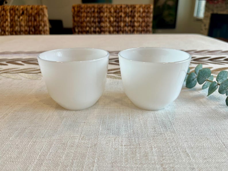 May include: Two white, opaque glass bowls with rounded bottoms and slightly flared rims. The bowls are set on a light-colored tablecloth with a subtle pattern. The background includes a woven chair and decorative elements.