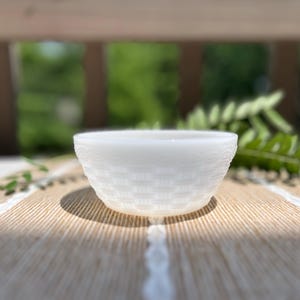 May include: A small, round, white milk glass bowl featuring a textured exterior pattern that mimics the appearance of a woven basket. The bowl has a smooth, slightly translucent rim and a consistent opaque white finish throughout its structure.