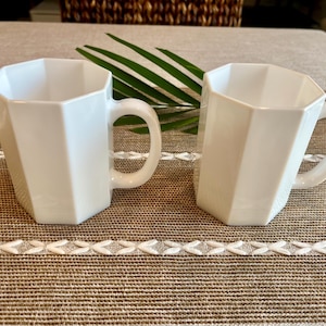 May include: Two white ceramic mugs with a geometric design. Each mug has a handle and a faceted exterior. The mugs are placed on a textured brown surface with a decorative white and beige trim. A green leaf is visible in the background.