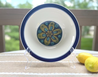 May include: A round ceramic bowl with a cream-coloured interior and a wide, dark blue rim. The centre of the bowl features a decorative floral pattern in shades of mustard yellow and teal blue, arranged in a symmetrical, petal-like design.