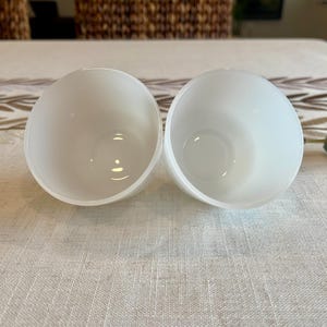 May include: Two white, semi-spherical bowls with a smooth, matte finish. The bowls are empty and have a rounded shape, with a slight inward curve at the top. They are placed side by side on a light-colored, textured surface.