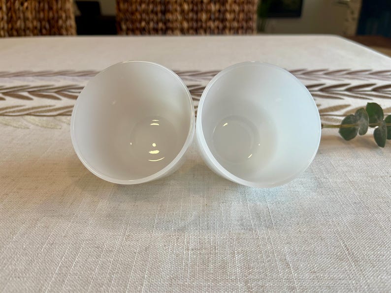 May include: Two white, semi-spherical bowls with a smooth, matte finish. The bowls are empty and have a rounded shape, with a slight inward curve at the top. They are placed side by side on a light-colored, textured surface.