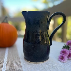 May include: A dark navy blue ceramic pitcher with a glossy, speckled finish, a curved handle, and a small pouring spout. The pitcher has a rounded body that tapers slightly toward the top and a visible light-colored base at the bottom edge.