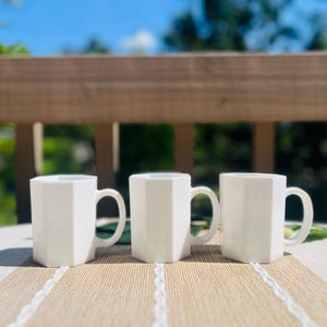 May include: Three white ceramic mugs with a distinct hexagonal shape and a smooth, glossy finish. Each mug features a sturdy, curved handle on the side, providing a consistent and uniform appearance across the set of three drinking vessels.