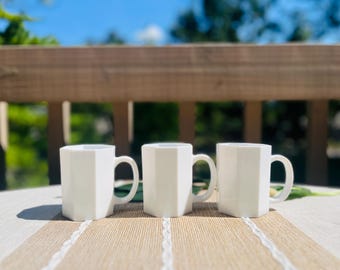 May include: Three white ceramic mugs with a distinct hexagonal shape and a smooth, glossy finish. Each mug features a sturdy, curved handle on the side, providing a consistent and uniform appearance across the set of three drinking vessels.