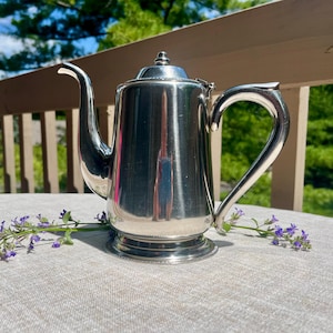 May include: A polished silver-toned metal teapot featuring a tall cylindrical body, a curved spout, a sturdy handle, and a small lid with a finial knob. The teapot rests on a circular base and is accented by small sprigs of purple flowers.