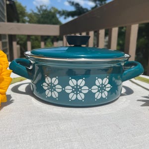 May include: A teal enamel pot with a matching lid and two side handles. The exterior features a white floral pattern consisting of three stylized flowers arranged in a horizontal row. The pot has a smooth, glossy finish and a silver-toned rim.