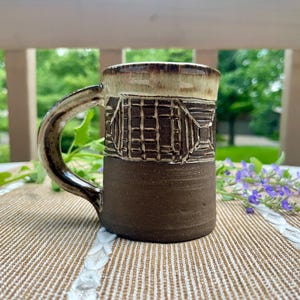 May include: A ceramic mug featuring a dark brown base and a textured upper section with a grid pattern. The rim and handle are finished in a glossy cream and brown glaze. The mug has a cylindrical body and a curved handle on the left side.