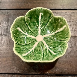 May include: A ceramic bowl shaped like a cabbage leaf, with a glossy green finish and white veins. The bowl is centered on a dark wooden surface. The cabbage leaf design is detailed and realistic.