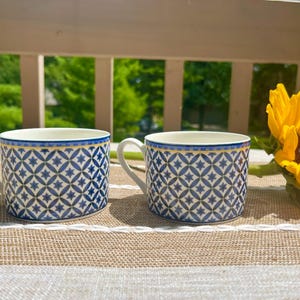 May include: Two ceramic cups featuring a repeating blue and white geometric star pattern with a thin gold rim. Each cup has a curved handle and a smooth white interior. The cups are positioned side by side on a textured tan and white surface.
