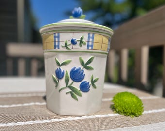 May include: A white ceramic canister with a lid featuring a blue tulip finial. The body is decorated with embossed blue tulips and green leaves. The upper rim has a yellow and blue checkered border with a thin green line accenting the lid edge.