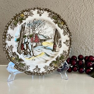 May include: A decorative plate with a winter scene featuring a red barn, snow-covered ground, and leafless trees. The plate has a scalloped edge with a brown and green floral design. The plate is displayed on a clear stand.