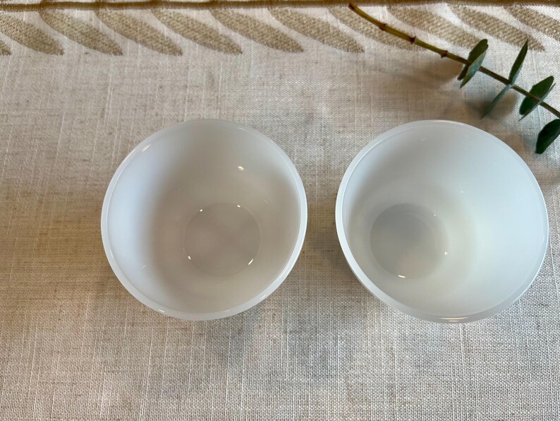 May include: Two white, frosted glass bowls are displayed on a neutral-toned, textured surface. The bowls are round with a simple, minimalist design. A sprig of greenery is visible in the upper right corner.
