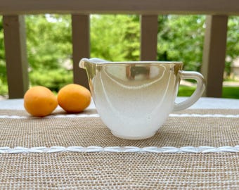 May include: A white milk glass creamer pitcher featuring a lustrous, iridescent gold finish on the upper portion, a curved pouring spout, and a small handle on the side, positioned next to two small, round, orange-coloured fruits.