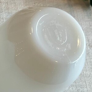 May include: A close-up of a white, opaque, rounded object, possibly a small bowl or cup. The object has a smooth, glossy surface with subtle imperfections. The bottom of the object has a slightly indented base with a faint marking. The background is a textured, light-colored fabric.
