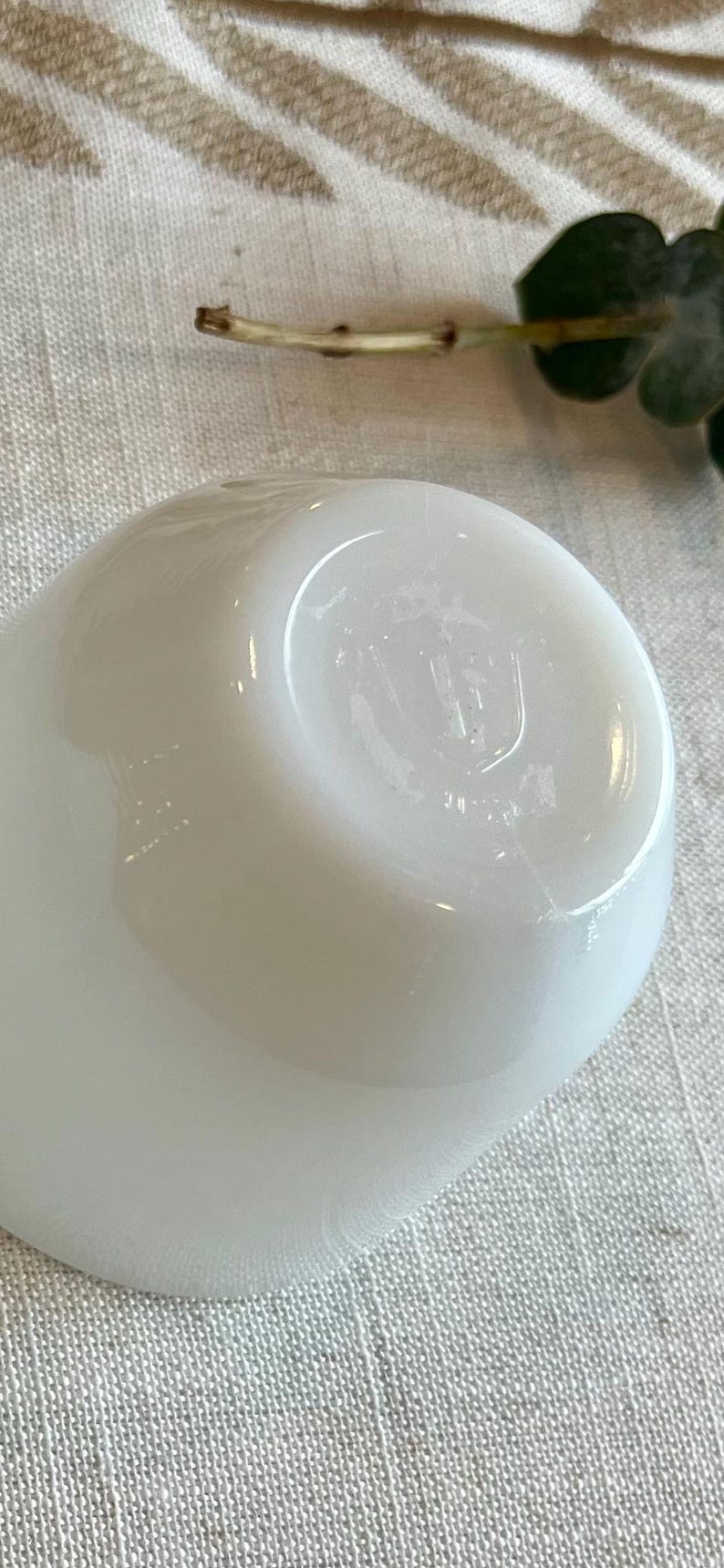 May include: A close-up of a white, opaque, rounded object, possibly a small bowl or cup. The object has a smooth, glossy surface with subtle imperfections. The bottom of the object has a slightly indented base with a faint marking. The background is a textured, light-colored fabric.