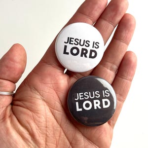 May include: Two round button pins with the text "JESUS IS LORD" in black lettering on a white background and a black background.