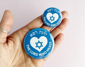 May include: Two blue round buttons with white text and a white heart containing a Star of David. The text reads "יהוה רפא" and "THE LORD WHO HEALS". The buttons are different sizes.