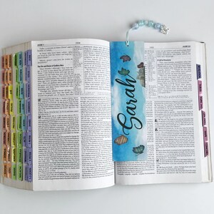 May include: An open book with colorful tabs and a bookmark. The bookmark features the name "Sarah" in cursive on a blue watercolor background with decorative elements. A beaded charm with a butterfly hangs from the bookmark.