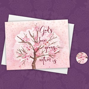 May include: A greeting card with a watercolor illustration of a flowering tree in shades of pink and brown. The text "God's love grows within us" is written in a cursive font. A matching round sticker is also visible.
