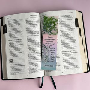 Puede incluir: Un primer plano de una Biblia abierta con un marcapáginas con un diseño floral y el texto "For the commandment is a lamp, The teaching is a light, And the way to life is the rebuke that disciplines. (Proverbs 6:23)"