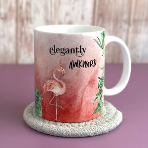 May include: White ceramic mug with a pink flamingo and green foliage design. The mug has the text "elegantly awkward" printed on it.