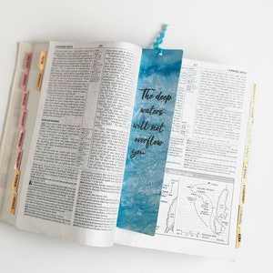 Könnte beinhalten: Ein blaues und weiß gemustertes Lesezeichen mit dem Text "The deep waters will not overflow you" in schwarzen Buchstaben. Das Lesezeichen befindet sich in einer geöffneten Bibel.
