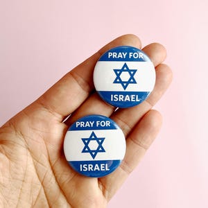 Puede incluir: Dos pines de botón azul y blanco con la Estrella de David y el texto "Pray for Israel".