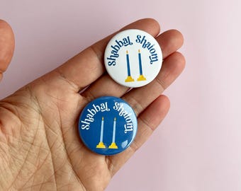 May include: Two round buttons with the text "Shabbat Shalom" and a graphic of two blue candles with gold bases. One button is white with blue text and candles, the other is blue with white text and candles.