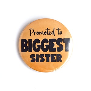 May include: A round, orange button with the text "Promoted to BIGGEST SISTER" in black, bold letters. The button is centered on a white background.