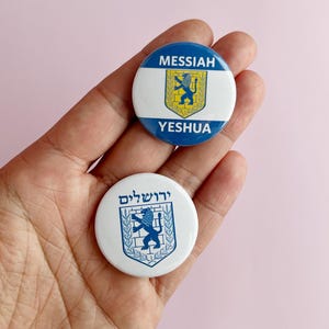 Peut inclure: Deux boutons ronds blancs et bleus avec un lion et un bouclier. Le bouton du haut dit "MESSIAH YESHUA" et le bouton du bas dit "ירושלים".