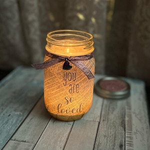 May include: A glass jar candle with a brown paper label that says "You are so loved". The jar is decorated with a purple ribbon and a small black heart.