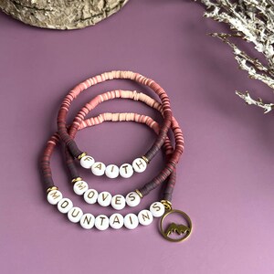 May include: Three layered bracelets with pink, red, and white beads. The beads spell out the phrase "Faith Moves Mountains" and a small mountain charm hangs from the bottom.