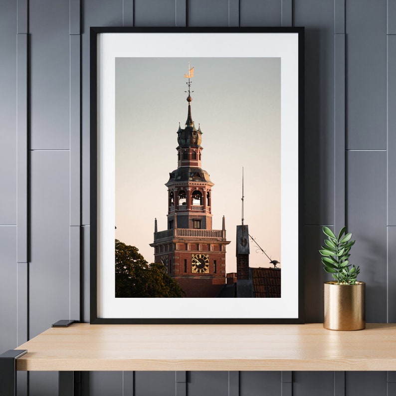 Pode incluir: Uma fotografia emoldurada de uma alta torre de tijolo vermelho com um mostrador de rel&oacute;gio e um telhado pontudo. A torre &eacute; cercada por &aacute;rvores e o c&eacute;u &eacute; azul claro.