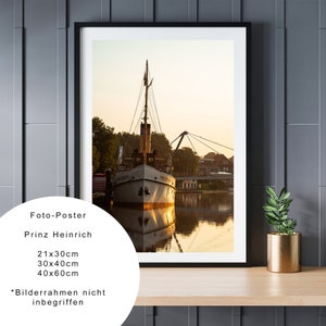 Pode incluir: Uma fotografia emoldurada de um navio branco atracado em um cais com uma ponte ao fundo. A água está calma e reflete a luz dourada do pôr do sol. O texto "Prinz Heinrich" é visível abaixo da imagem.