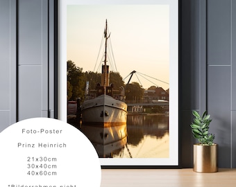 Puede incluir: Una fotografía enmarcada de un barco blanco atracado en un muelle con un puente en el fondo. El agua está tranquila y refleja la luz dorada de la puesta de sol. El texto "Prinz Heinrich" es visible debajo de la imagen.