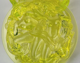 May include: A translucent, yellow-green glass lid and base. The lid is a shallow bowl with a ribbed design. The base is a circular shape with a detailed relief of a classical figure. The glass has a slight glow.
