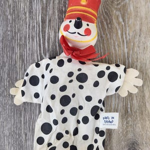 May include: A vintage hand puppet featuring a clown-like face with a red hat and bow tie. The puppet's shirt is white with black polka dots and a red trim. The tag reads "BABES IN TOYLAND".
