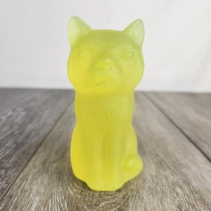 May include: A translucent yellow cat figurine. The cat is sitting upright with its ears perked up. The figurine has a smooth texture and is made of a material that allows light to pass through. The cat is sitting on a wooden surface.