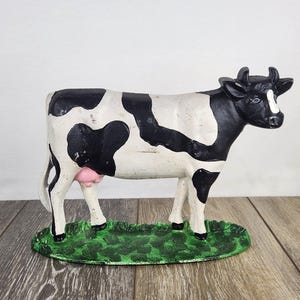 May include: A cast iron cow figurine, painted in black and white spots, stands on a green base. The cow has a pink udder and black horns. The figurine is a decorative item.