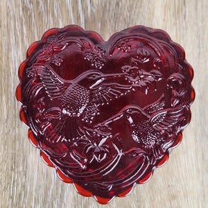 May include: A heart-shaped, ruby-red glass trinket box. The box features an embossed design of hummingbirds and floral elements. The edges have a scalloped design. The box is likely used for storing small jewelry items or keepsakes.