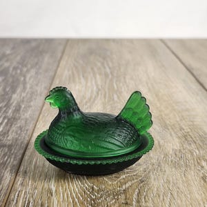 May include: A translucent emerald green glass hen on a nest dish. The lid is shaped like a sitting hen with detailed feather textures, while the base features a beaded rim design. The glass has a smooth, matte finish throughout the piece.