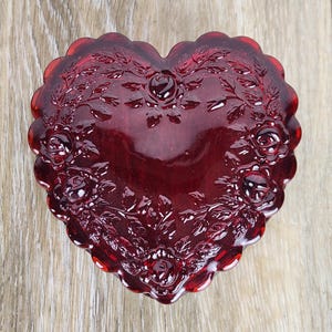 May include: A heart-shaped, ruby-red glass trinket box. The box features a detailed floral design with roses and leaves. The edges are scalloped, and the box is sitting on a light wood surface.