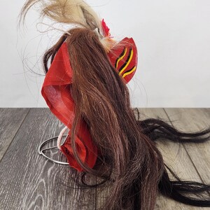Pu&ograve; includere: Una tradizionale maschera Barong indonesiana, con tessuto rosso vibrante, lunghi capelli castani e neri e accenti decorativi. La maschera ha un design stilizzato con dettagli gialli e neri e una base in filo di ferro.