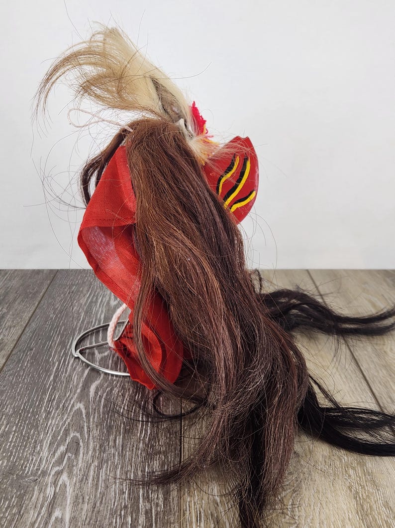 Pu&ograve; includere: Una tradizionale maschera Barong indonesiana, con tessuto rosso vibrante, lunghi capelli castani e neri e accenti decorativi. La maschera ha un design stilizzato con dettagli gialli e neri e una base in filo di ferro.