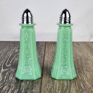 May include: Two matching green glass salt and pepper shakers with silver-colored metal tops. The shakers have a floral design and are displayed on a wooden surface. The shakers are a light green color.
