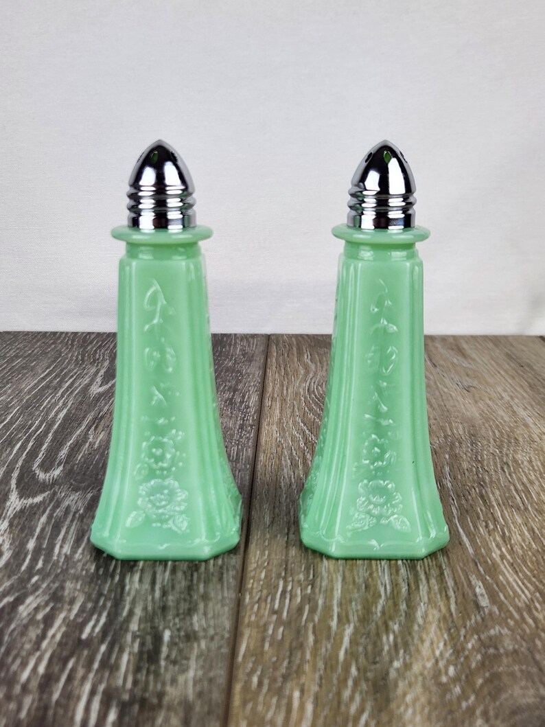 May include: Two matching green glass salt and pepper shakers with silver-colored metal tops. The shakers have a floral design and are displayed on a wooden surface. The shakers are a light green color.