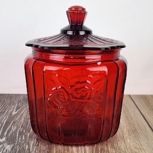 May include: A ruby red glass jar with a lid. The jar has a floral design and a faceted lid. The jar is sitting on a wooden surface. The jar is a vintage style.