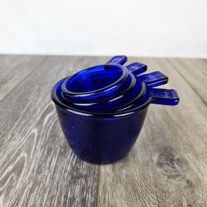 May include: A set of four blue glass measuring cups, nested together. The cups are made of cobalt blue glass and have a smooth, rounded shape.